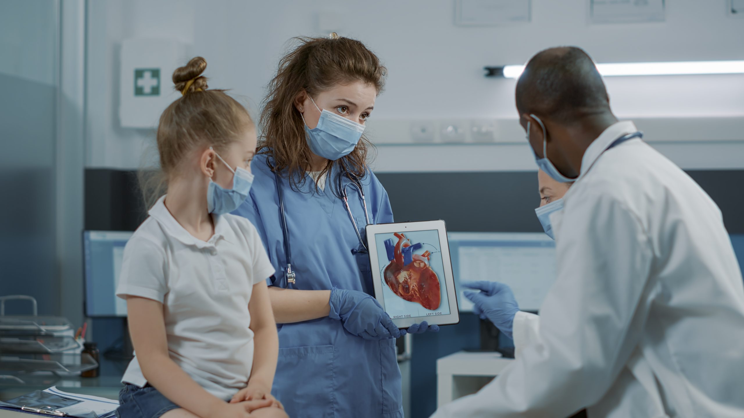 Woman nurse showing cardiology image on digital tablet, letting doctor explain cardiovascular diagnosis to child and parent. Assistant holding modern device with heart anatomy picture.